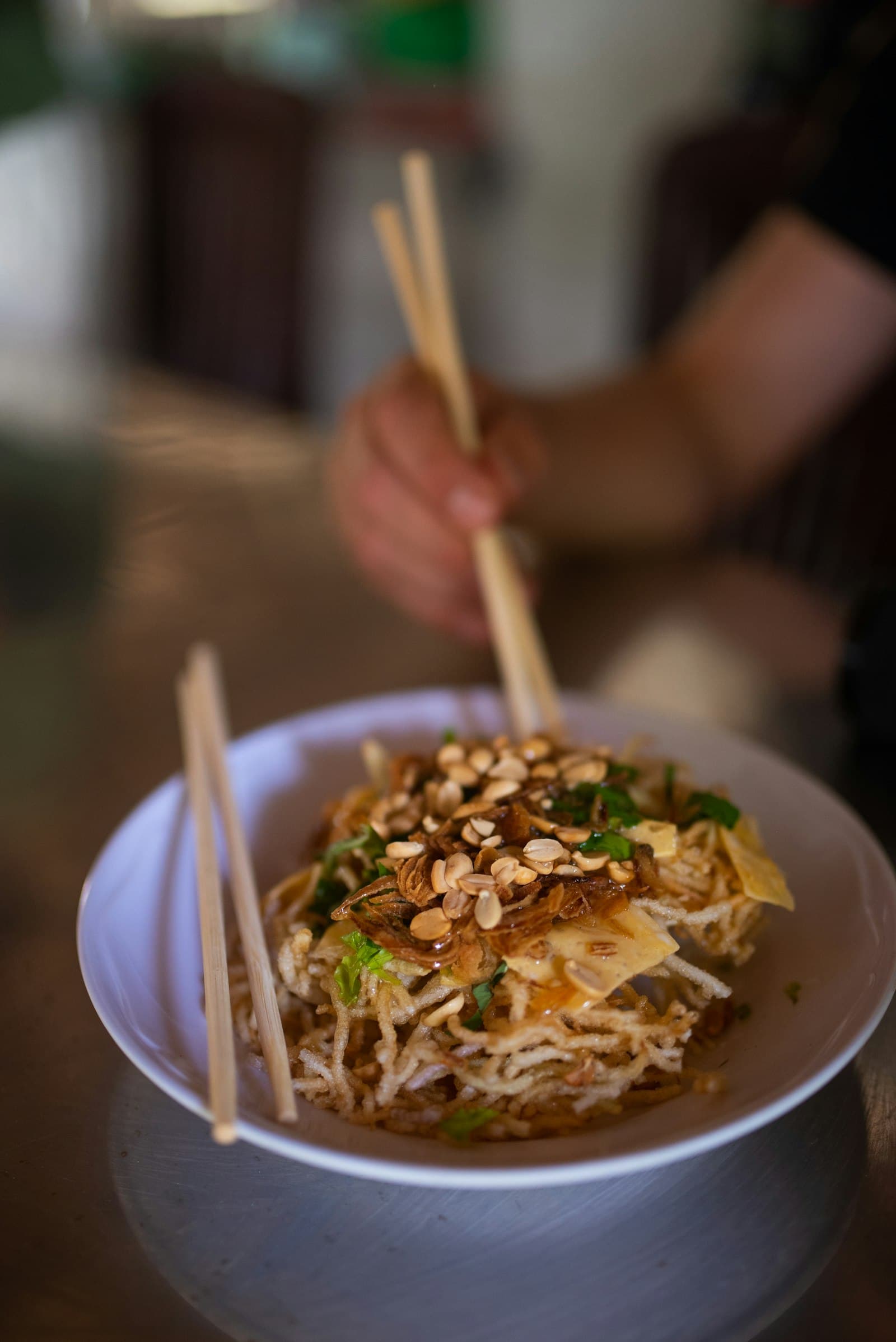 A bowl of noodles topped with chopped peanuts and herbs, with chopsticks